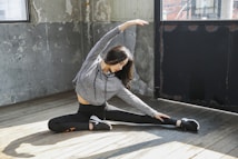 A person is seated on a wooden floor in a spacious room doing a side stretch. They are wearing a gray hoodie and black leggings with athletic shoes. The walls have a rustic, worn appearance with peeling paint, and there are large windows allowing natural light to illuminate the space.