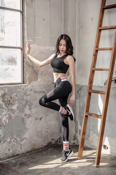 A woman in athletic wear is standing in a sunlit, industrial-style room beside a wooden ladder. She is leaning against a worn, textured wall with one leg raised, wearing black leggings, a black sports bra, and sneakers with striped socks. Natural light pours in through the large window, casting soft shadows.