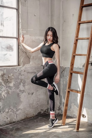 A woman in athletic wear is standing in a sunlit, industrial-style room beside a wooden ladder. She is leaning against a worn, textured wall with one leg raised, wearing black leggings, a black sports bra, and sneakers with striped socks. Natural light pours in through the large window, casting soft shadows.