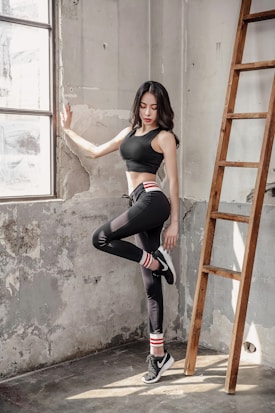 A woman in athletic wear is standing in a sunlit, industrial-style room beside a wooden ladder. She is leaning against a worn, textured wall with one leg raised, wearing black leggings, a black sports bra, and sneakers with striped socks. Natural light pours in through the large window, casting soft shadows.