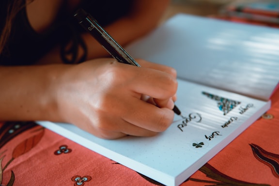 A hand holding a black pen writing words on a white notebook, which is resting on a red tablecloth with floral patterns. The notebook has a drawing of a Christmas tree and some text.