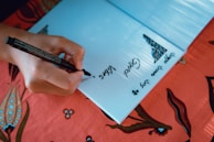 A hand is writing in a notebook with a black marker. The notebook is open on a table covered with a colorful textile that has floral patterns. There are words written on the page, including 'Good vibes'.