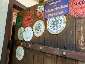 A wooden door displaying several circular and shield-shaped plaques, each with various inscriptions and symbols related to agricultural awards or affiliations. The plaques vary in color, including red, green, white, and purple, and are attached with visible screws. The door has visible iron hinges and is part of a rustic structure, with a portion of the white exterior wall shown.