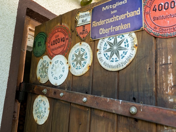 A wooden door displaying several circular and shield-shaped plaques, each with various inscriptions and symbols related to agricultural awards or affiliations. The plaques vary in color, including red, green, white, and purple, and are attached with visible screws. The door has visible iron hinges and is part of a rustic structure, with a portion of the white exterior wall shown.