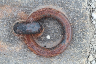 A close-up view of a rusted metal ring embedded in a rough concrete surface. The ring shows signs of oxidation with an uneven, textured appearance. There are small pieces of gravel scattered around on the concrete.