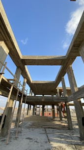 An unfinished concrete building with a skeletal framework, showing multiple floors and columns under a clear blue sky. Wooden planks are scattered on the ground, and ladders are positioned against the structure.
