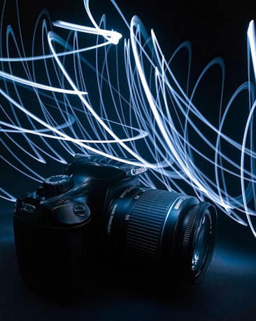 A DSLR camera is positioned against a dark background with swirling light trails. The camera's lens and buttons are visible, and the light trails create dynamic patterns of motion around it.