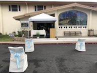 A church entrance with an outdoor setup featuring a canopy tent over a table and a wooden podium. The area is decorated with white chairs adorned with light blue ribbons, facing the podium. There's a large stained-glass window on the building façade and several benches lined up beneath it.