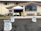 A church entrance with an outdoor setup featuring a canopy tent over a table and a wooden podium. The area is decorated with white chairs adorned with light blue ribbons, facing the podium. There's a large stained-glass window on the building façade and several benches lined up beneath it.
