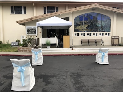 A church entrance with an outdoor setup featuring a canopy tent over a table and a wooden podium. The area is decorated with white chairs adorned with light blue ribbons, facing the podium. There's a large stained-glass window on the building façade and several benches lined up beneath it.