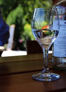 A clear wine glass filled with water is placed on a wooden table. The glass has some text printed on it. In the background, there is a blurred view of greenery and sunlight, indicating an outdoor setting. A bottle partially appears to the right of the glass.