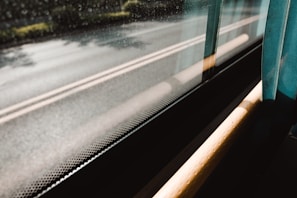 Sunlight casts shadows on the interior of a vehicle as it moves past a blurred street view. The window is slightly fogged or has condensation, suggesting cool or rainy weather outside. A teal curtain partially covers the window, blending with the reflections. The warm light creates an inviting atmosphere.