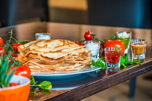 A large stack of thin, golden-brown pancakes is placed on a white plate. Surrounding the pancakes are small glass containers holding various sauces and dips, including a red jam and what appears to be a white cream. Fresh tomatoes, green herbs, and leafy greens are placed around the plate, adding vibrant color and freshness to the scene.