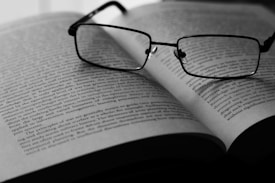 A pair of eyeglasses rests on an open book, with text visible on the pages. The image is in black and white, highlighting the contrast between the dark frames of the glasses and the lighter background of the book. The focus appears to be on the glasses, with the text slightly blurred in the background.