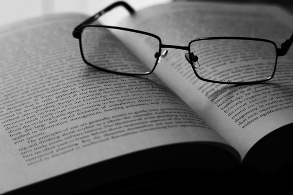 A pair of eyeglasses rests on an open book, with text visible on the pages. The image is in black and white, highlighting the contrast between the dark frames of the glasses and the lighter background of the book. The focus appears to be on the glasses, with the text slightly blurred in the background.