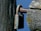 A close-up view of a weathered lock attached to a rusty metal bar. The lock and bar are affixed to an old wooden post, showing signs of age and exposure to the elements with moss and lichen growth. The background is a clear blue sky.