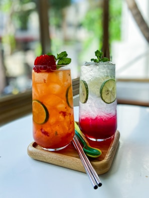 Two tall, chilled drinks are presented on a wooden tray. The left drink is a vibrant orange with visible lime slices and a strawberry garnish. The right drink displays a gradient from clear to deep pink, featuring lime slices and topped with mint leaves. The scene includes metal straws and a light background that suggests a sunny setting.