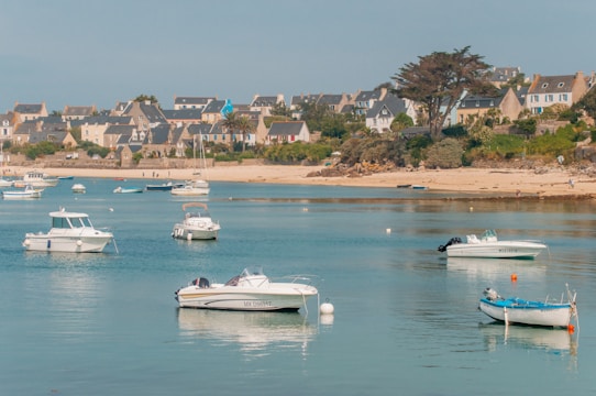 Several small boats float on calm blue waters near a sandy shoreline. In the background, there is a picturesque village with stone houses and colorful shutters, surrounded by greenery and trees.