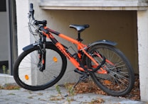 A bright orange and black mountain bike is parked against a wall. The bike features thick tires and a sturdy frame, suitable for off-road cycling. The ground is covered with some dry leaves and small patches of grass, and the scene gives a casual, everyday vibe.