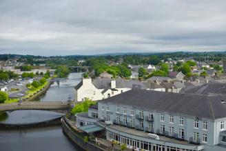 A scenic view of a quaint town with a river running through it. The town features a collection of buildings with the River Court Hotel prominently displayed in the foreground. A bridge spans the river, connecting different parts of the town, while lush green trees and vegetation are scattered throughout the landscape. The sky is overcast, suggesting mild weather, and the overall atmosphere appears calm and picturesque.