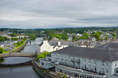 A scenic view of a quaint town with a river running through it. The town features a collection of buildings with the River Court Hotel prominently displayed in the foreground. A bridge spans the river, connecting different parts of the town, while lush green trees and vegetation are scattered throughout the landscape. The sky is overcast, suggesting mild weather, and the overall atmosphere appears calm and picturesque.