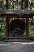 A wooden structure featuring a large cross-section of a tree trunk with visible growth rings is placed on a wooden deck. The structure has a thatched roof made of twigs and branches. Surrounding the structure is a dense forest with tall trees and lush greenery.