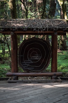 A wooden structure featuring a large cross-section of a tree trunk with visible growth rings is placed on a wooden deck. The structure has a thatched roof made of twigs and branches. Surrounding the structure is a dense forest with tall trees and lush greenery.
