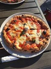 A freshly baked pizza with a crispy, slightly charred crust sits on a white plate. It is topped with melted mozzarella cheese, vibrant red tomato sauce, and fresh basil leaves. The pizza is placed on a wooden table, and there is a hint of sunlight illuminating parts of the crust.