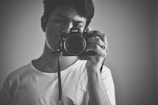 A person takes a self-portrait using a camera. The black and white image captures the intense and focused expression of the individual, who is dressed in a casual white T-shirt. The closeness of the shot emphasizes the details of the camera and the person's face.