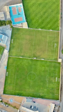 An aerial view of two adjacent green grass soccer fields with white goalposts and markings. Nearby, there is a blue tennis court and surrounding infrastructure including buildings and parked cars.