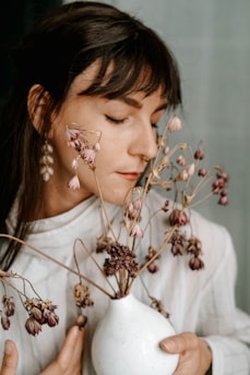A woman with closed eyes gently holds a white vase with dried flowers. She appears to be inhaling their scent or embracing their presence, dressed in a soft, light-colored outfit. The overall setting is calm and serene.