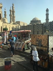A street market scene featuring several stalls displaying various goods, including jewelry and souvenirs. Two individuals are seen browsing the merchandise. In the background, historic Islamic architecture with tall minarets and a dome can be observed, suggesting a culturally rich area.