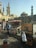 A street market scene featuring several stalls displaying various goods, including jewelry and souvenirs. Two individuals are seen browsing the merchandise. In the background, historic Islamic architecture with tall minarets and a dome can be observed, suggesting a culturally rich area.