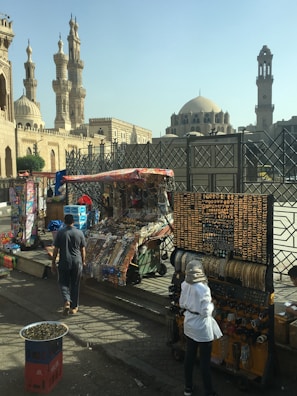 A street market scene featuring several stalls displaying various goods, including jewelry and souvenirs. Two individuals are seen browsing the merchandise. In the background, historic Islamic architecture with tall minarets and a dome can be observed, suggesting a culturally rich area.
