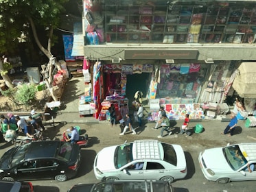 A bustling street scene depicts a marketplace with people walking along the sidewalk. Various textiles are displayed outside a shop, with bright colors seen on the hanging fabric and stacked goods. Cars, including a taxi, are parked and driving along the street. The atmosphere is busy with vendors and pedestrians.