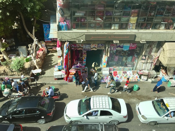 A bustling street scene depicts a marketplace with people walking along the sidewalk. Various textiles are displayed outside a shop, with bright colors seen on the hanging fabric and stacked goods. Cars, including a taxi, are parked and driving along the street. The atmosphere is busy with vendors and pedestrians.