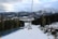 A ski slope surrounded by tall trees, with ski lifts ascending up the mountain. Snow-covered mountains are visible in the background, under a partly cloudy sky. Small groups of people can be seen skiing down the slope.