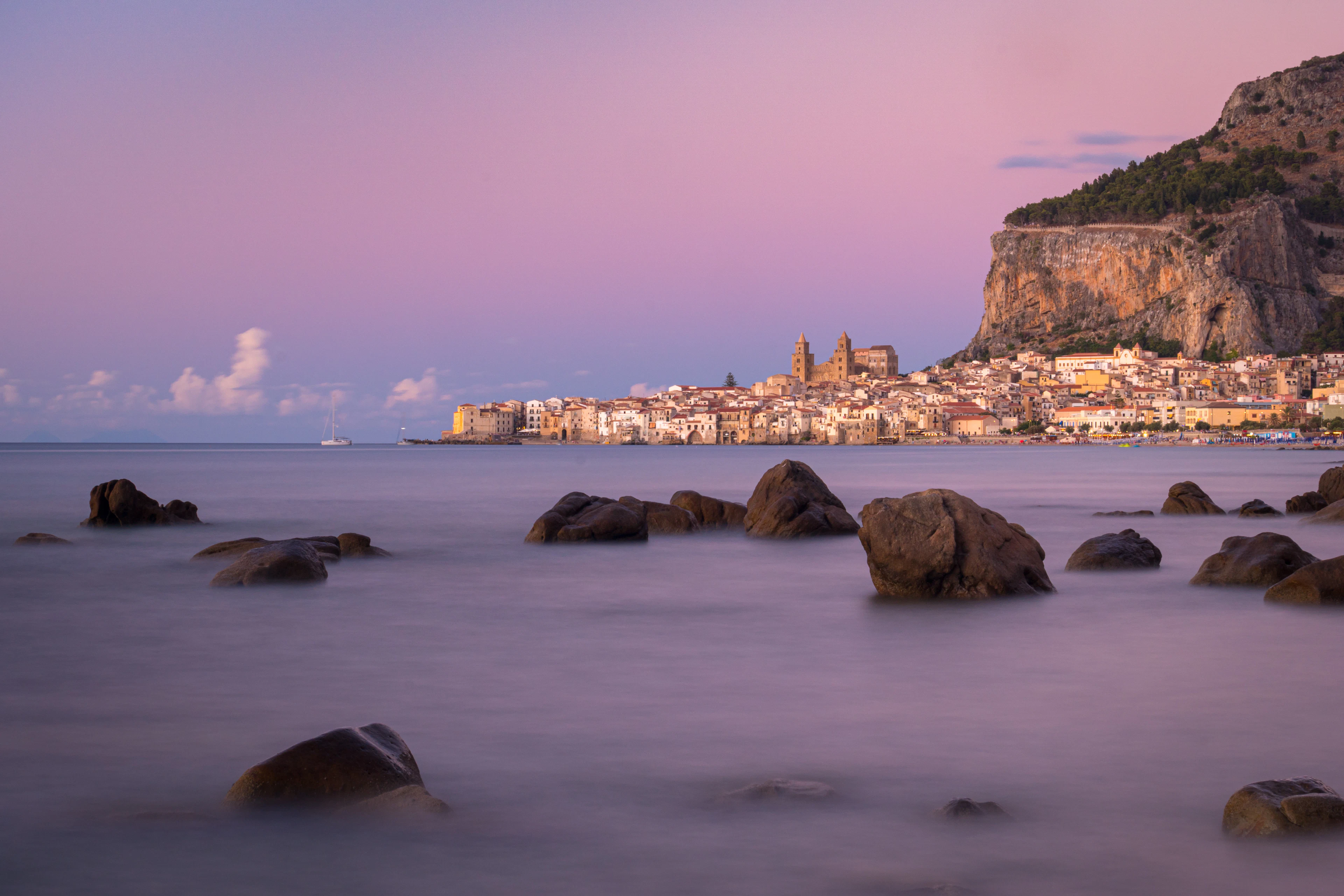 Cefalù senamiestis ir katedra po Rocca uola mėlynąją valandą, pirmame plane – akmenys ramioje jūroje