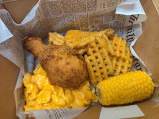 A box lined with newspaper print contains a meal consisting of a fried chicken drumstick, waffle-cut fries, macaroni and cheese, and a cob of corn.