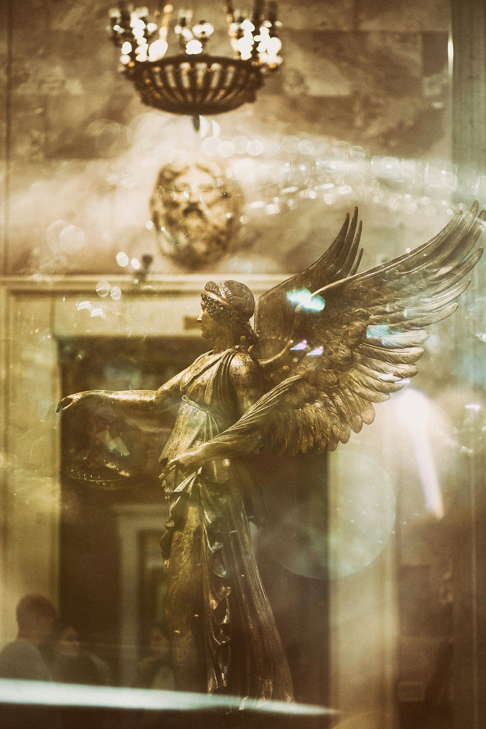A bronze statue of an angel with large wings is depicted, set in a dimly lit room with ornate decor. A chandelier hangs from the ceiling, and reflections and bokeh effects add a dreamlike quality to the scene. A carved face adorns the wall in the background, adding to the intricate and historical ambiance.