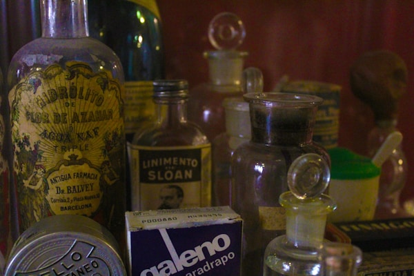 A collection of vintage glass bottles and containers, some labeled with old-style typography. The bottles have ornate designs, and labels feature text like 'Hidrolito' and 'Linimento Sloan'. The scene evokes a nostalgic or historic mood, reminiscent of an old apothecary or pharmacy setting.