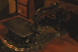 An old, vintage sewing machine with intricate, ornate details is situated on a wooden surface. The machine has a metal body with decorative, faded gold accents, and appears to be aged. Towards the back, a small wooden chest with a drawer is partially visible.