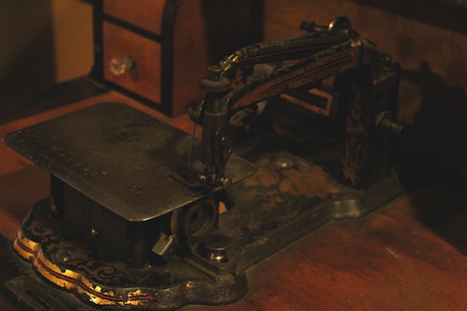 An old, vintage sewing machine with intricate, ornate details is situated on a wooden surface. The machine has a metal body with decorative, faded gold accents, and appears to be aged. Towards the back, a small wooden chest with a drawer is partially visible.