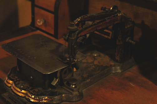 An old, vintage sewing machine with intricate, ornate details is situated on a wooden surface. The machine has a metal body with decorative, faded gold accents, and appears to be aged. Towards the back, a small wooden chest with a drawer is partially visible.