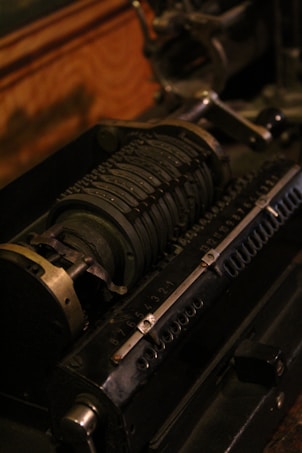 An antique mechanical calculator with a series of rotating cylindrical drums marked with numbers. It features metal levers and gears, showcasing intricate engineering. The device appears to be aged, with a dark, industrial aesthetic.