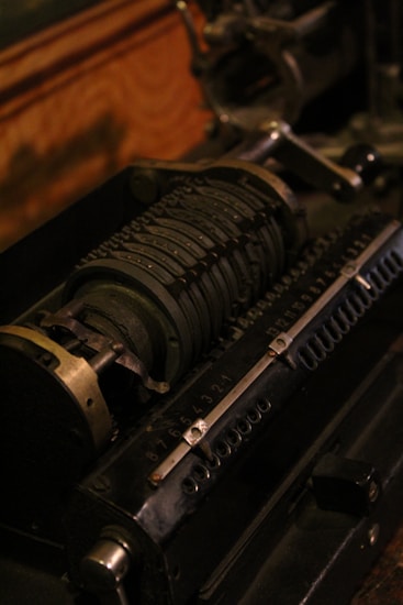 An antique mechanical calculator with a series of rotating cylindrical drums marked with numbers. It features metal levers and gears, showcasing intricate engineering. The device appears to be aged, with a dark, industrial aesthetic.