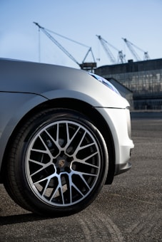 The image captures a close-up of a car's front wheel with sleek alloy rims. In the background, there are several large industrial cranes and a warehouse-like building, indicating an industrial or port setting. The lighting suggests an early morning or late afternoon setting.