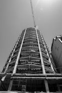 A black and white photograph of a tall building under construction, featuring a steel framework and concrete columns. A crane extends from the top, reaching out into the sky, with sunlight filtering through.