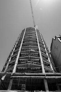 A black and white photograph of a tall building under construction, featuring a steel framework and concrete columns. A crane extends from the top, reaching out into the sky, with sunlight filtering through.