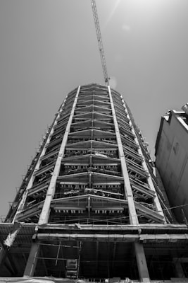 A black and white photograph of a tall building under construction, featuring a steel framework and concrete columns. A crane extends from the top, reaching out into the sky, with sunlight filtering through.
